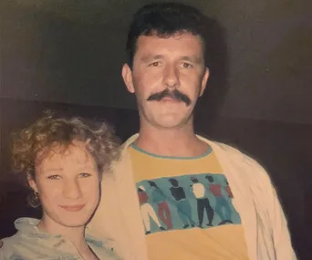 A man with a mustache and a woman with curly hair smiling, captured in a casual setting.