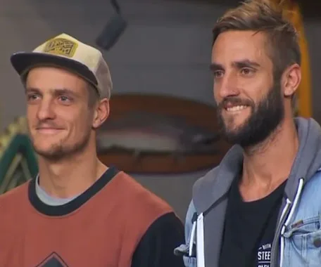 Two men standing side by side, one wearing a cap and sweater, the other in a jacket with a beard, both smiling.