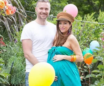 A couple poses outdoors; the woman is pregnant, wearing a green dress and hat, surrounded by colorful balloons.