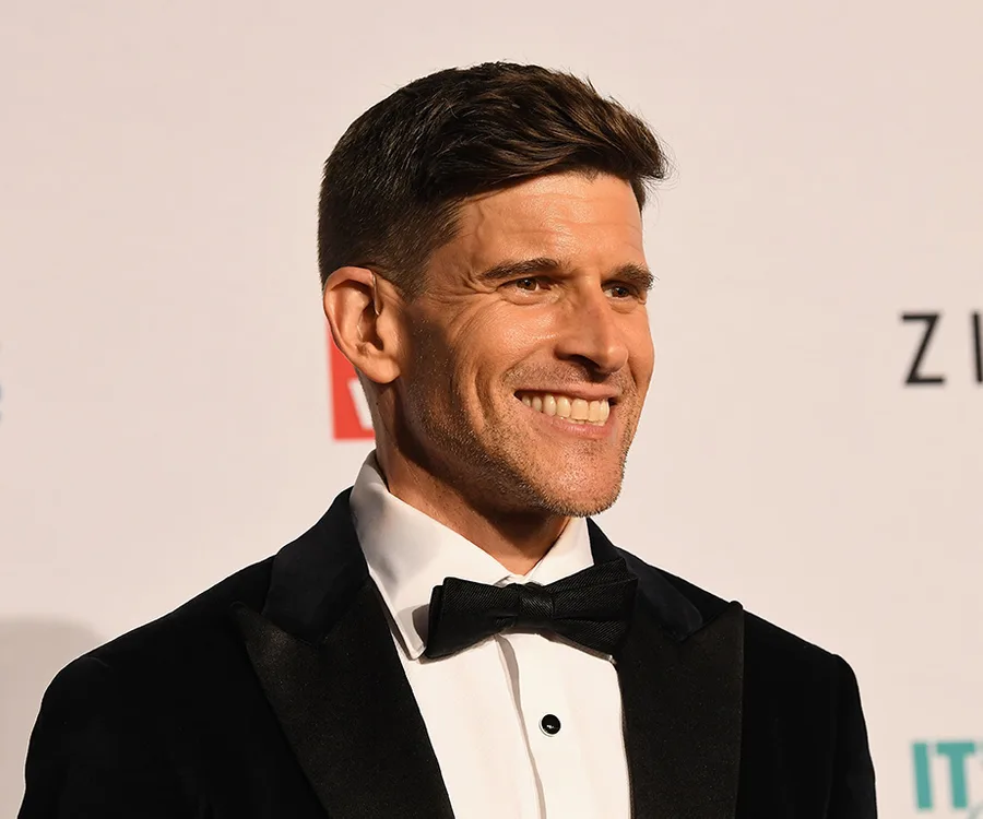 A person in a black tuxedo and bow tie smiles on a formal event's red carpet.
