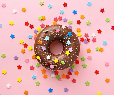 A chocolate donut with colorful star-shaped sprinkles on a pink background, surrounded by scattered star candies.