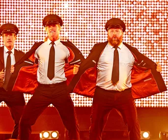 Male performers in suits, opening jackets, wearing pilot hats, on a brightly lit stage during a dance routine.