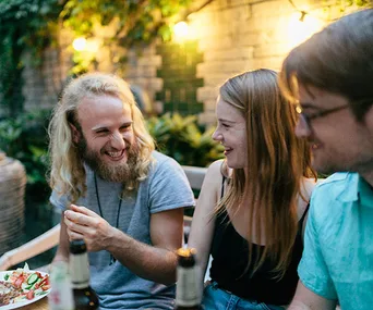 A group of three people laughing and enjoying drinks outdoors, illuminated by warm string lights.