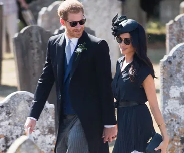 A couple dressed formally walks hand in hand, wearing sunglasses at an outdoor event, with gravestones in the background.
