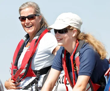 Two women smiling and wearing life jackets, sunglasses, sailing on a sunny day.