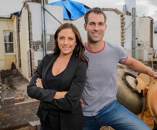 A smiling man and woman pose in front of a partially constructed building site.