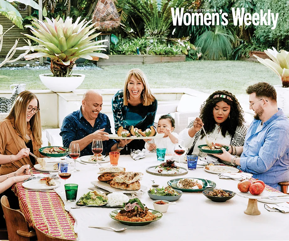 Group enjoying a vibrant outdoor meal with various dishes on a long table, under lush greenery in a garden setting.
