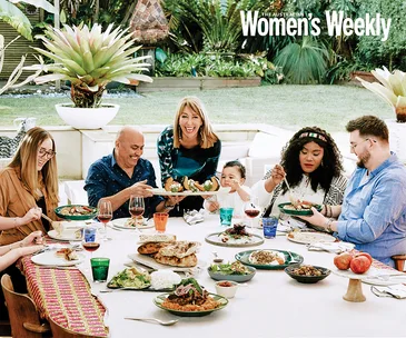 Group enjoying a vibrant outdoor meal with various dishes on a long table, under lush greenery in a garden setting.
