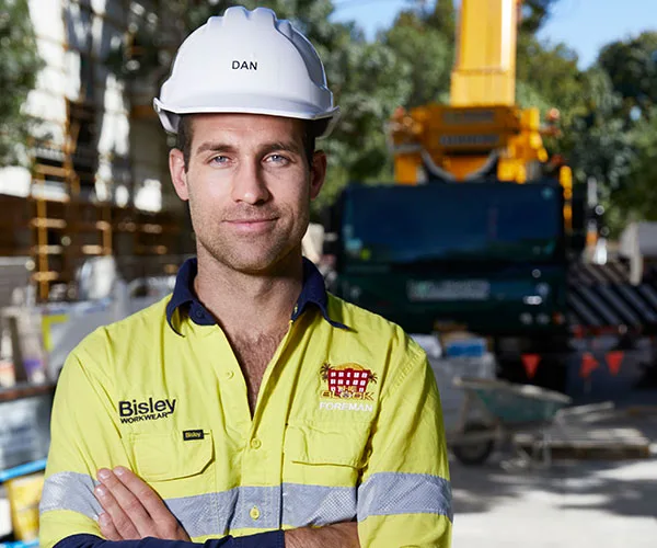 Foreman wearing a white hard hat labeled "Dan" and a yellow safety shirt with "The Block" logo, standing on a construction site.