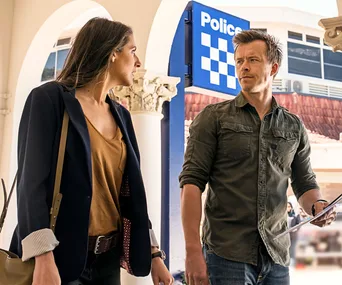 Two individuals converse outside a police station, with one holding documents and a visible police sign in the background.