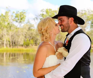 A bride and groom embrace by a pond, the groom in a cowboy hat and the bride holding a bouquet, looking fondly at each other.