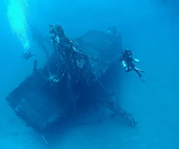 Divers exploring a sunken shipwreck on the ocean floor, with marine life beginning to cover the structure.