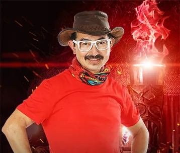 Man in a red shirt, glasses, and hat, stands confidently with tribal torch in the background on a Survivor set.