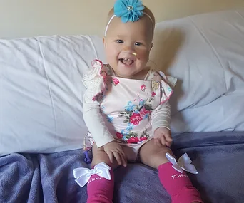 Smiling baby in floral outfit, pink socks with bows, and a turquoise headband sits on bed.