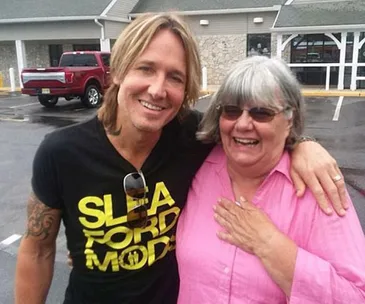 Man with long hair and band shirt smiles, arm around older woman in pink, outside a building.