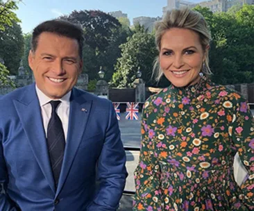 Two people smiling outdoors, with greenery and Union Jack flags in the background.