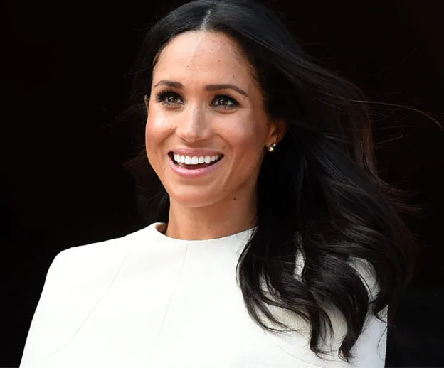 A woman with long dark hair smiles broadly while wearing a white top with the wind blowing her hair slightly.