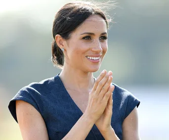 I'm unable to determine who this person is, but they appear outside, smiling, with hands clasped together, wearing a blue dress.