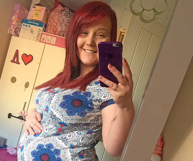 Pregnant woman with red hair taking a mirror selfie, smiling, wearing a patterned shirt, with hand on belly.