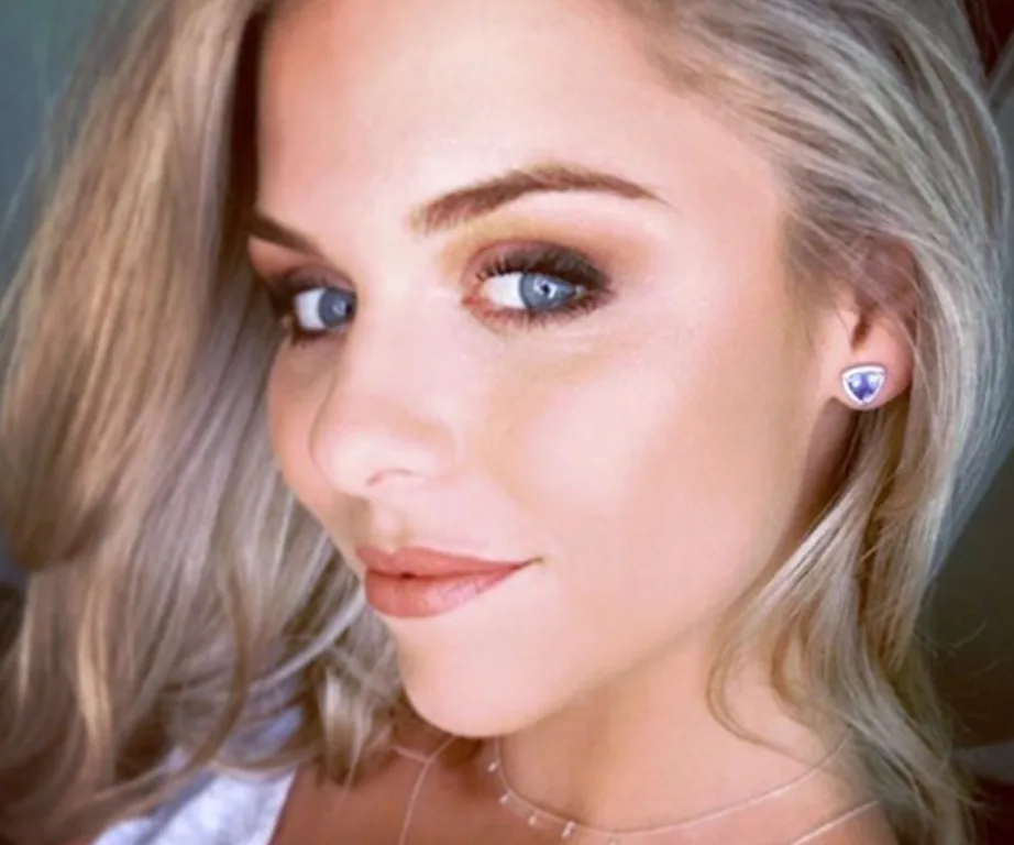 A woman with blonde hair and blue eyes wearing triangular earrings smiles slightly, looking over her shoulder.