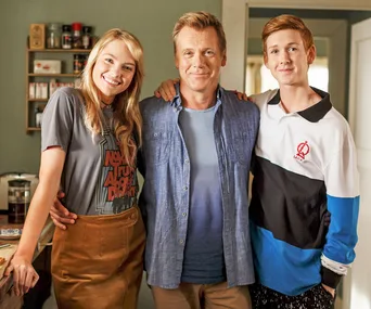 A cheerful trio stands together inside a cozy, warmly-lit room, smiling at the camera.