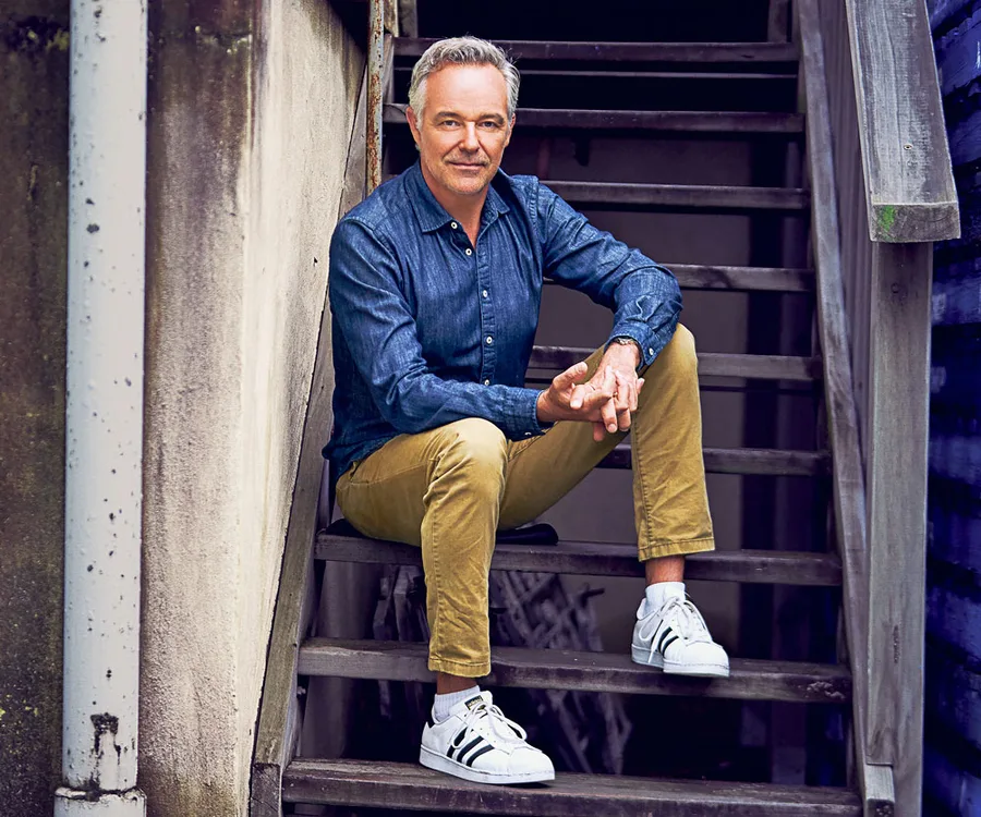 Man seated on outdoor stairs, wearing a blue shirt, beige pants, and white sneakers, with a relaxed expression.
