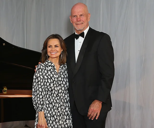 Couple posing together in formal attire, woman in patterned dress and man in black tuxedo, standing indoors.