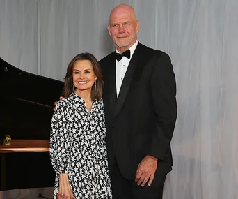 Couple posing together in formal attire, woman in patterned dress and man in black tuxedo, standing indoors.
