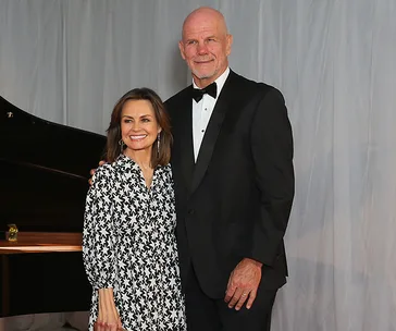 Couple posing together in formal attire, woman in patterned dress and man in black tuxedo, standing indoors.