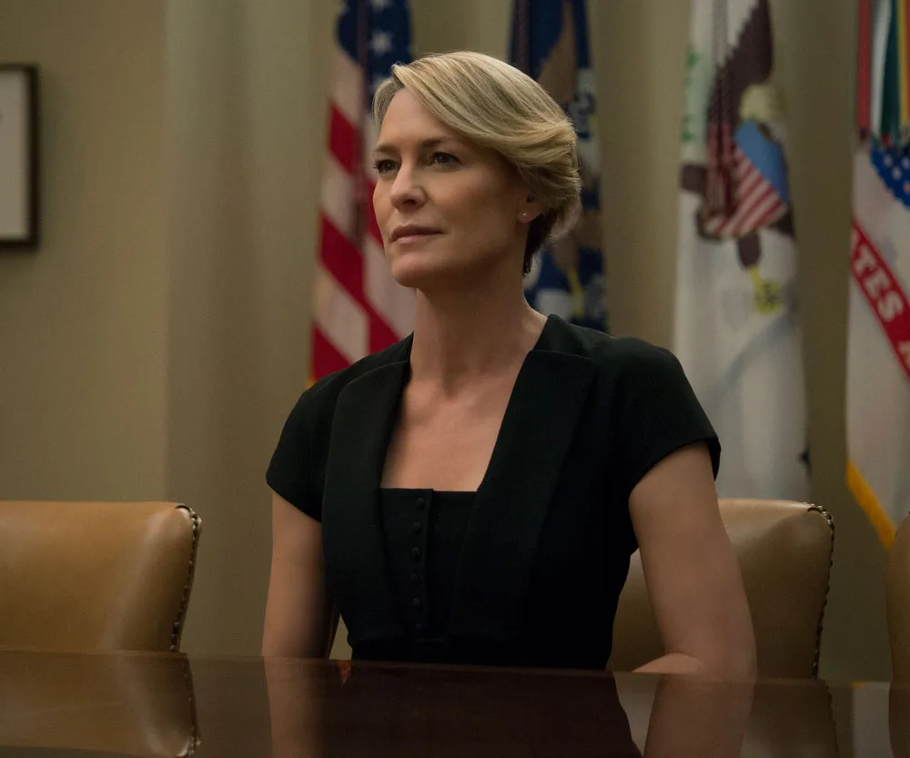 Claire Underwood from House of Cards sits at a conference table in a formal setting, with flags in the background.