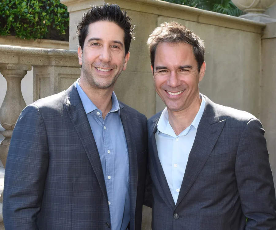 Two men in suits smiling, standing outdoors beside a stone banister.