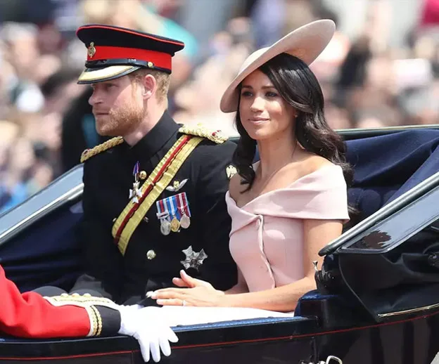 A couple dressed in formal attire rides in a carriage during a public event, with crowds visible in the background.