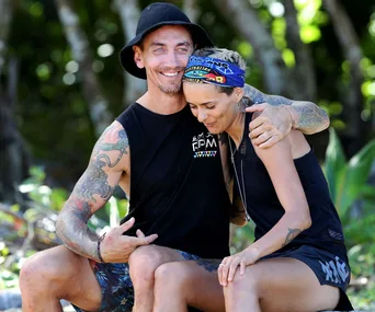 A man and woman sitting together outdoors, smiling and embracing; woman wearing an "Australian Survivor" bandana.