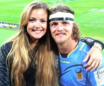 A man in a blue sports jersey with a headband poses on a field with a woman in a black jacket, both smiling.