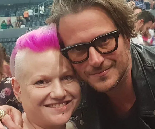 A man with glasses and a woman with pink hair smile together at an event.