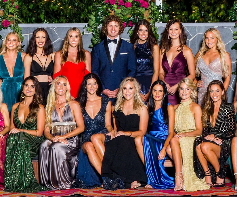 Group photo of Nick Cummins in a suit with 17 women in evening dresses, set against a floral backdrop.