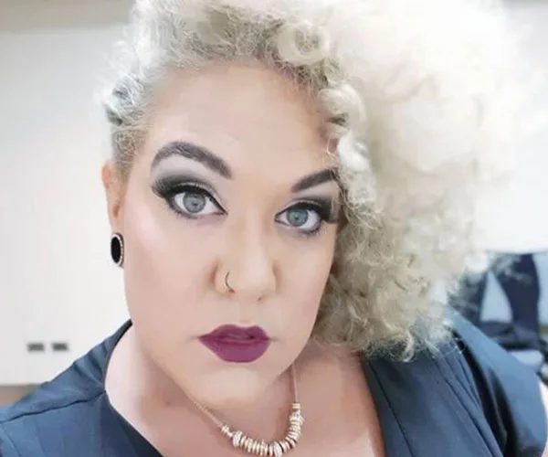 Female with curly blonde hair, wearing makeup, nose ring, earrings, and necklace, looking at the camera.