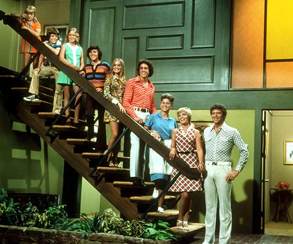 "The Brady Bunch family stands on a staircase in colorful 70s attire inside their iconic TV house."