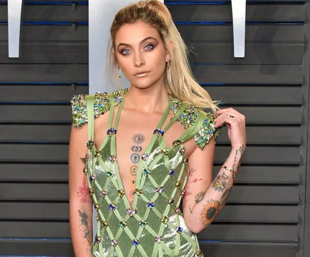 A woman in a green, embellished dress with tattoos poses on a stylized gray background.