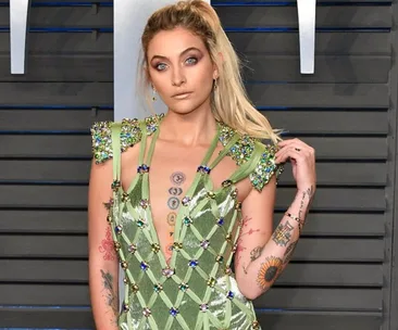 A woman in a green, embellished dress with tattoos poses on a stylized gray background.