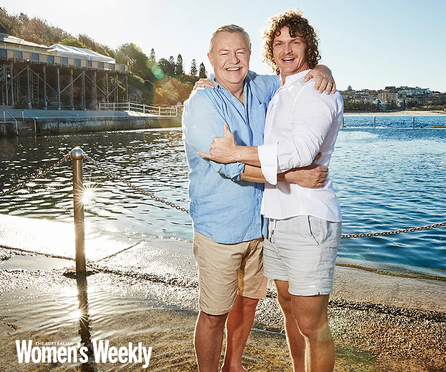 Two men smiling and embracing near a waterfront, under a clear blue sky.