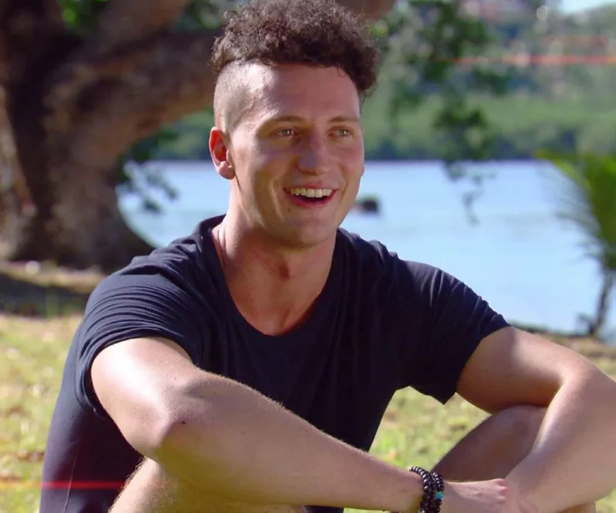 Young man sitting outdoors, smiling, with short curly hair, wearing a black t-shirt. Trees and water in the background.
