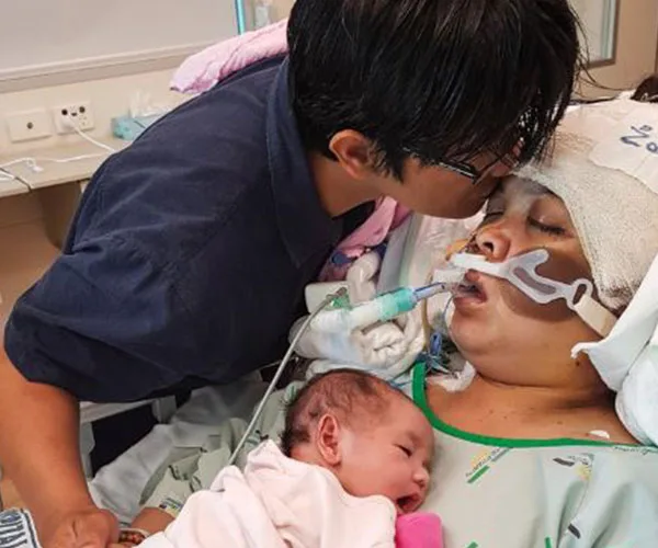 Son kisses mother on hospital bed; newborn sleeps nearby.