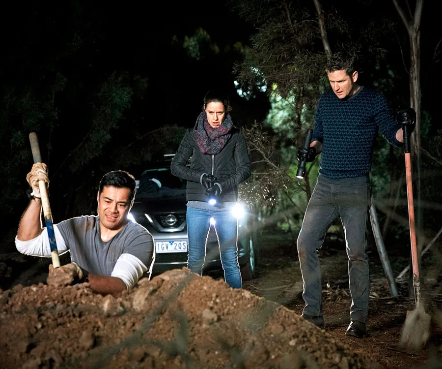 A man digging a hole at night, observed by a woman with a flashlight and another man holding a shovel, near a car.