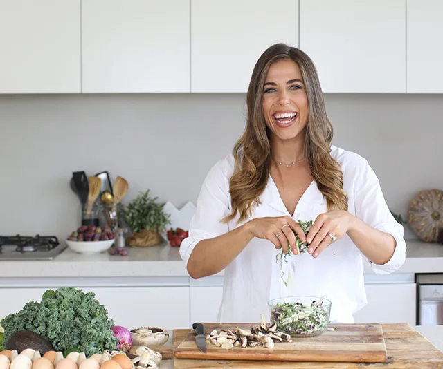 Person in a kitchen preparing a salad, surrounded by fresh vegetables, smiling at the camera.