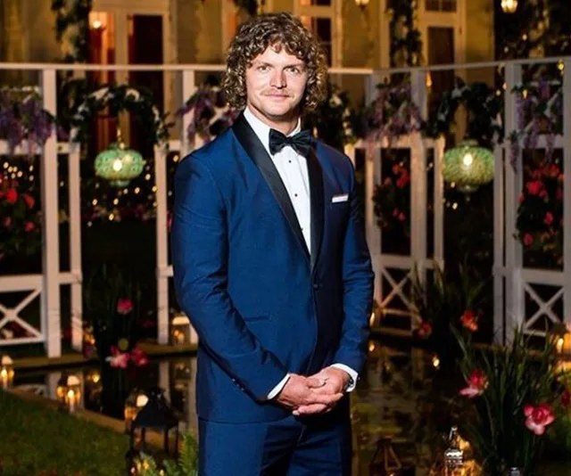 A man in a blue tuxedo stands in a decorated garden with lights and flowers at night.