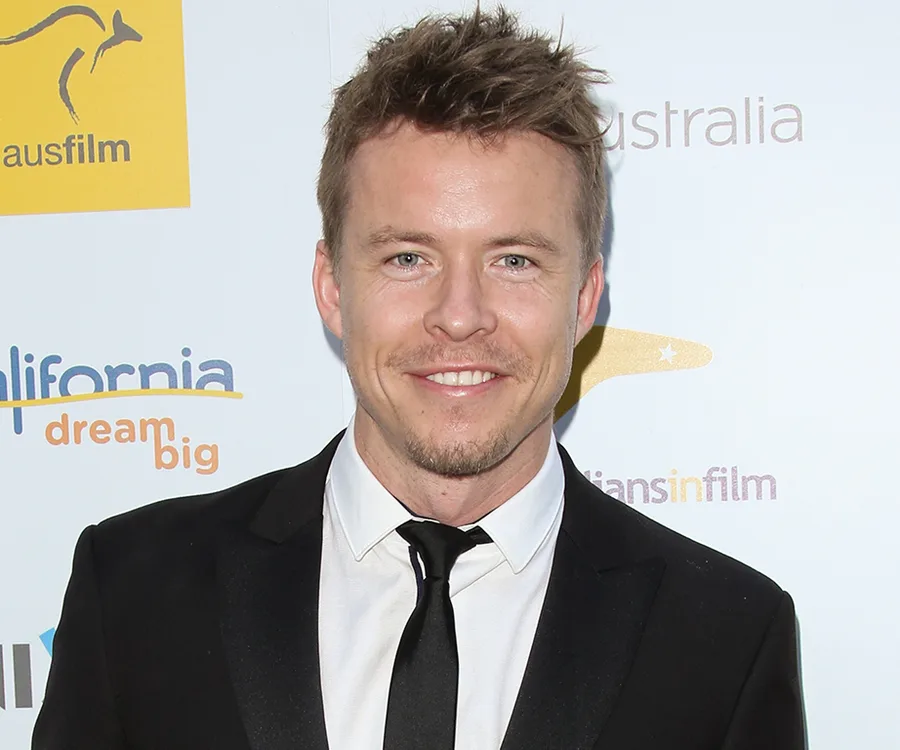 Celebrity in a formal suit at a promotional event, smiling at the camera, with logos in the background.