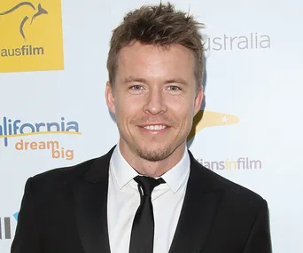Celebrity in a formal suit at a promotional event, smiling at the camera, with logos in the background.