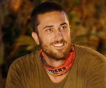 A contestant from "Survivor Australia" at tribal council wearing a red Survivors buff, smiling and looking to the side.