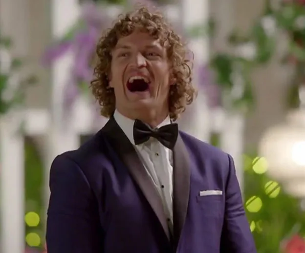 A man in a tuxedo laughs joyfully at a formal event with floral decor in the background.